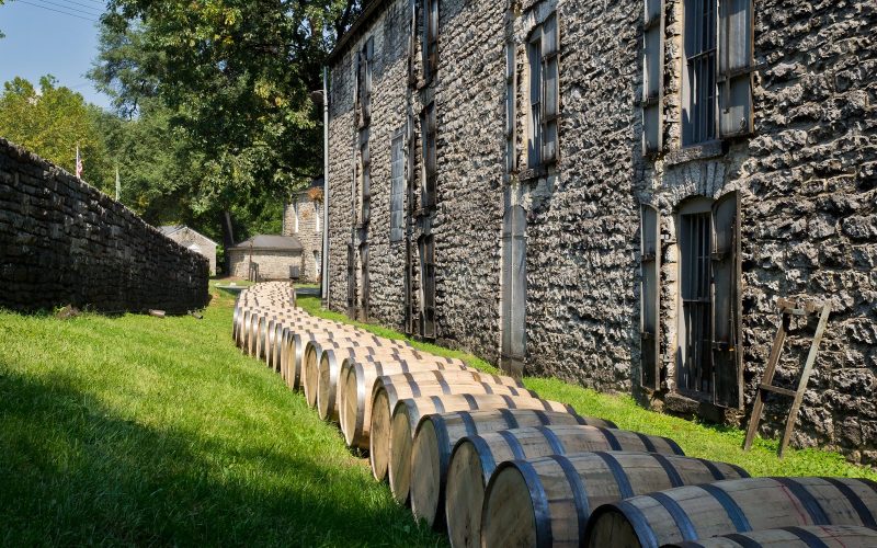 Bourbon barrels lined up on the grass beside a stone rickhouse at a Kentucky distillery