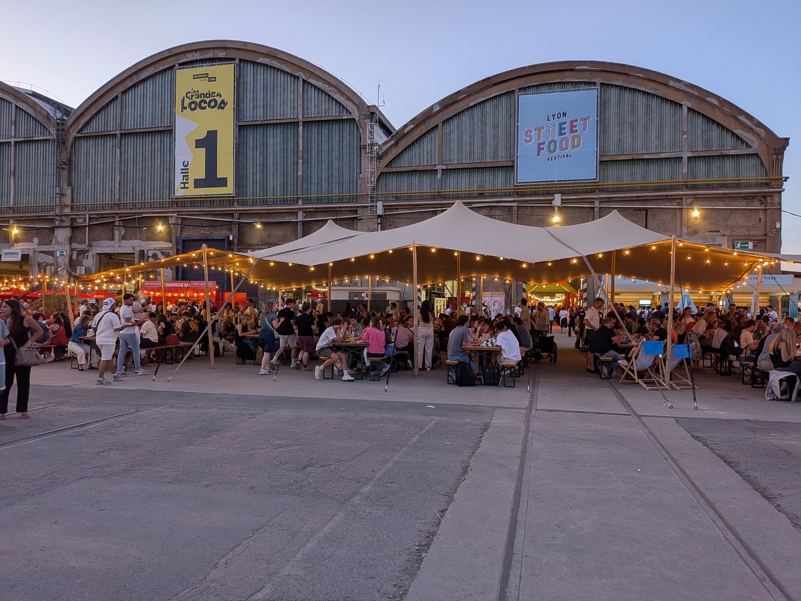 Diners on the central terrace under string lights in front of the Grandes Locos halls at dusk